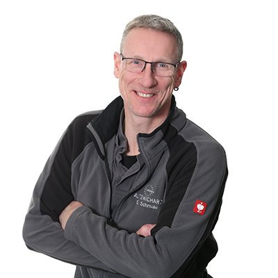 Ein freundlicher Mann mit Brille und kurzen Haaren steht lächelnd vor einer neutralen weißen Wand, die seine Persönlichkeit unterstreicht.