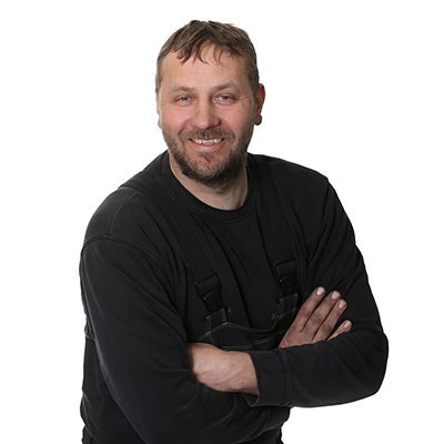 Ein lächelnder Mann mit Bart steht mit verschränkten Armen vor einem weißen Hintergrund, gekleidet in einem schwarzen Oberteil.