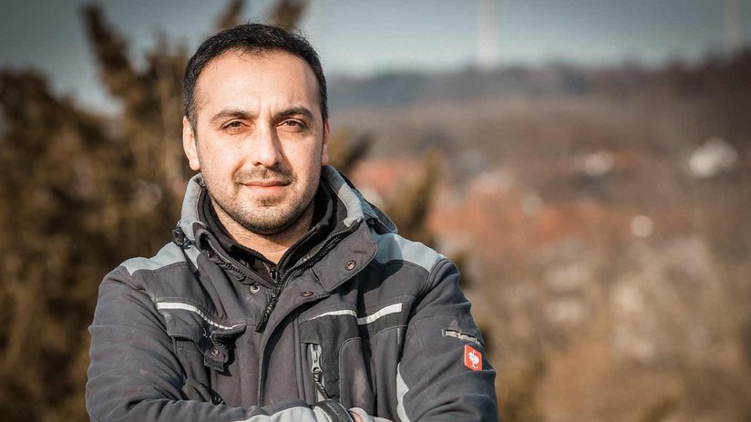 Ein Mann mit dunklem Haar und Bart steht selbstbewusst mit verschränkten Armen vor einer unscharfen Landschaft.