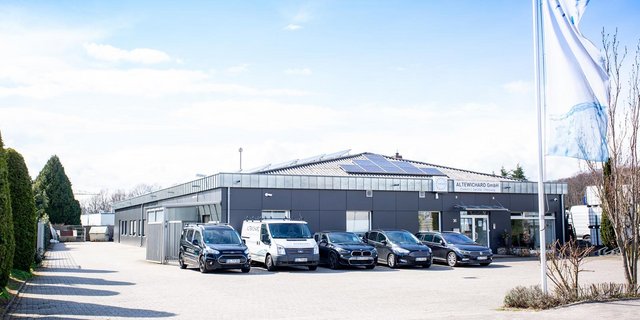 Ein modernes Firmengebäude mit Solarpanelen auf dem Dach, umgeben von mehreren Autos auf einem gepflasterten Parkplatz.