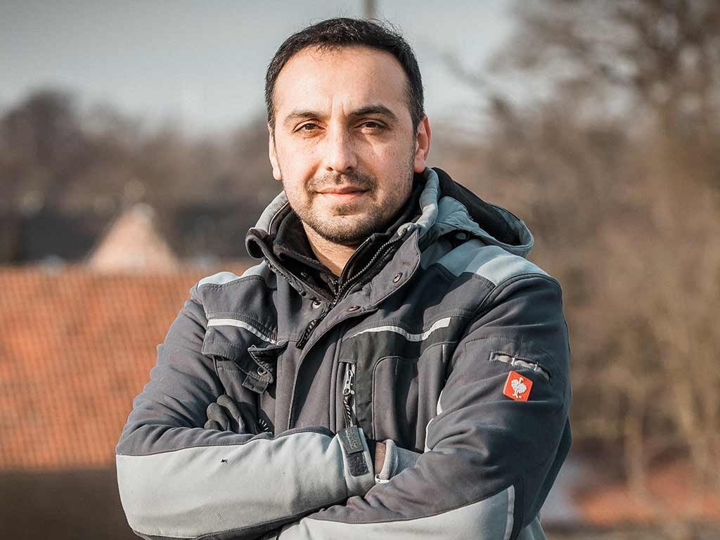 Ein Mann mit verschränkten Armen steht selbstbewusst im Freien, umgeben von einer unscharfen, natürlichen Kulisse.