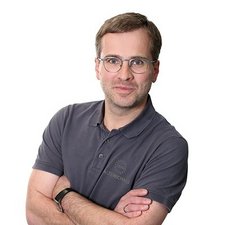 Professionelles Porträt eines Mannes mit Brille Der Mann wirkt freundlich und selbstsicher, während er in einem grauen Poloshirt mit Brille vor einem hellen Hintergrund steht.